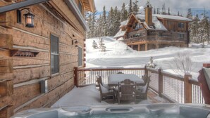 A wooden cabin with a hot tub and outdoor seating, surrounded by snow-covered trees and a ski slope. - Big Sky Vacation Rentals: PR Manitou 5 (Gallatin Gateway)