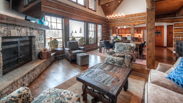 A spacious living room with a stone fireplace, wooden beams, and large windows offering views of the outdoors. - Big Sky Vacation Rentals: PR Manitou 5 (Gallatin Gateway)
