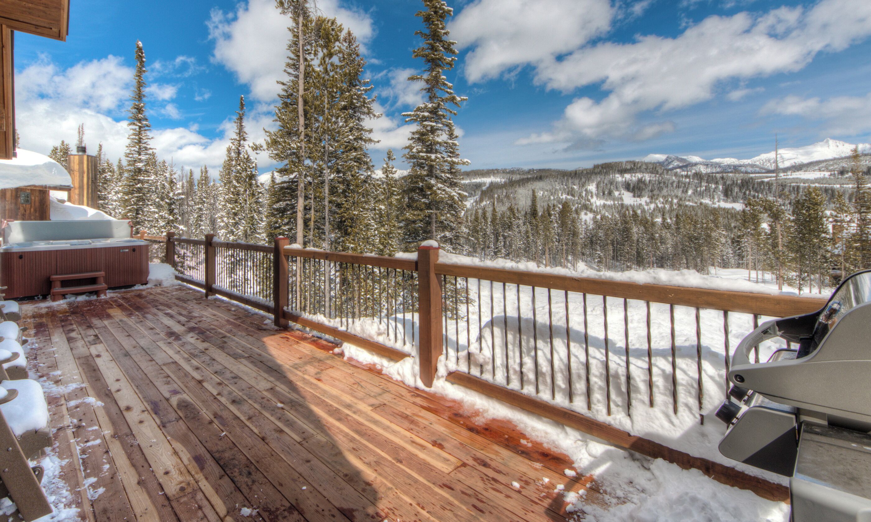 A wooden deck with a hot tub and grill, overlooking a snowy mountain landscape.