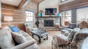 A cozy living room with a stone fireplace, wooden beams, and a large TV. - Big Sky Vacation Rentals: PR Manitou 5 (Gallatin Gateway)