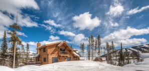 A wooden cabin with a balcony, surrounded by snow-covered ground and tall pine trees, set against a backdrop of mountains and a partly cloudy sky. - Big Sky Vacation Rentals: PR Manitou 5 (Gallatin Gateway)