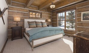 A log cabin bedroom with a bed, bedside lamps, a window with a view of snow-covered trees, and a painting on the wall. - Big Sky Vacation Rentals: PR Manitou 5 (Gallatin Gateway)