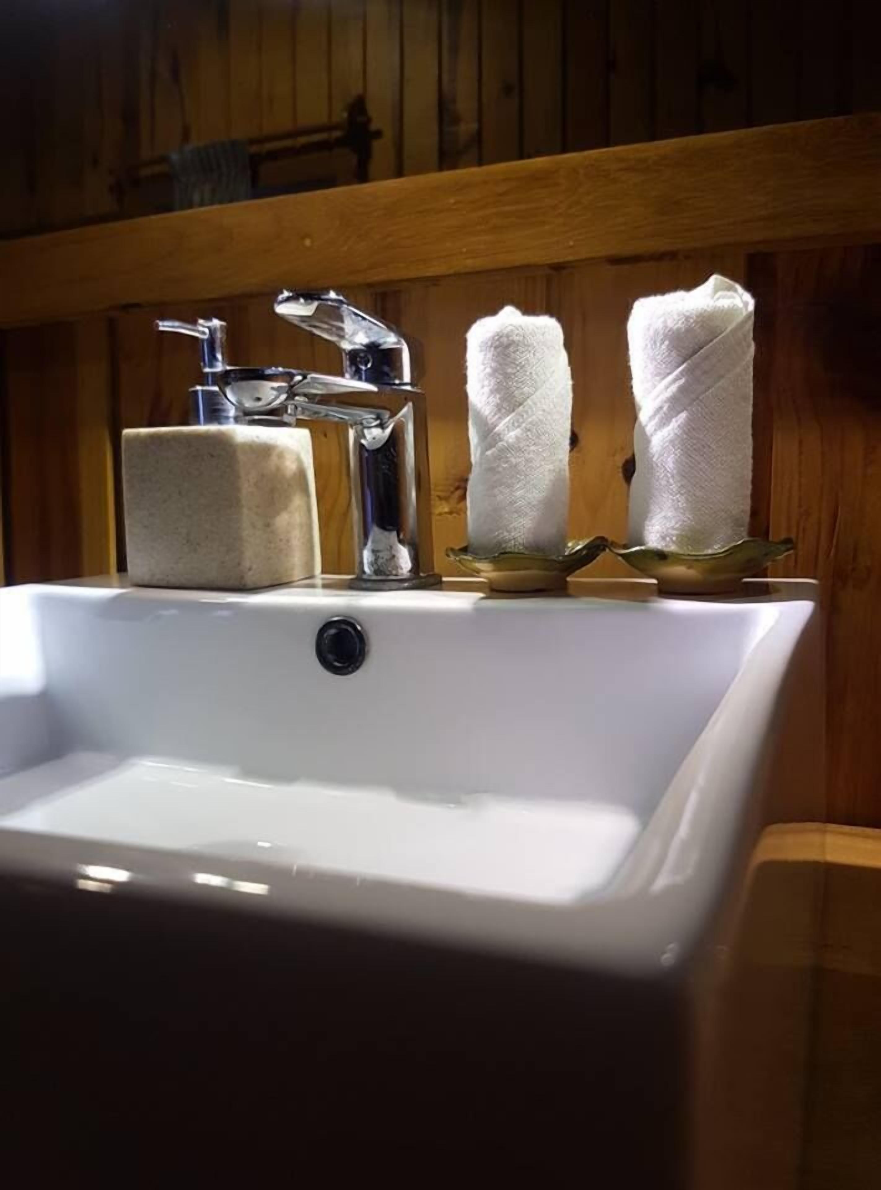 Un lavabo con grifo cromado, dos toallas blancas sobre un estante de madera y un jabonero.