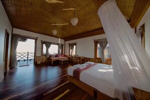 Une pièce spacieuse avec un grand lit, un plancher en bois et un plafond haut surmonté d’un toit de chaume. Elle est dotée de fenêtres avec des rideaux, d’un balcon et offre une vue sur la mer.