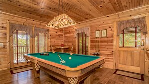 Un intérieur en bois avec une table de billard, un luminaire suspendu et des fenêtres équipées de stores.