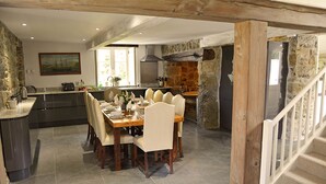 Une salle à manger spacieuse, avec une grande table en bois dressée pour six personnes, entourée de chaises. On aperçoit, au fond, une cuisine avec un mur en pierre et un four moderne.