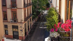 A narrow street with buildings on either side, featuring balconies with railings and potted plants.