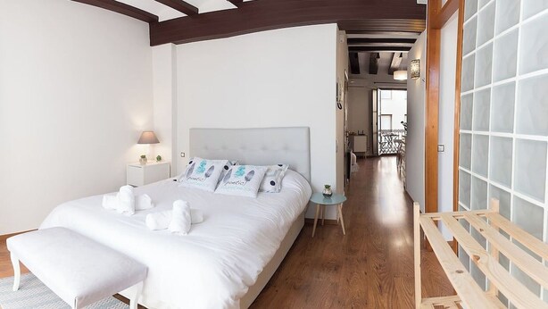 A bedroom with a large bed, bedside tables, a nightstand, a wooden bench, and a view of a balcony.