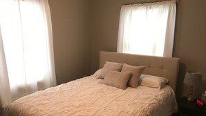 Una cama bien hecha con una colcha color beige y almohadas, una lámpara de noche y una mesita de noche con un reloj.