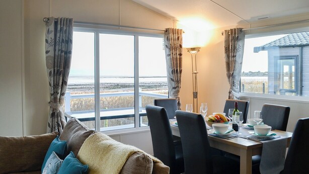 A dining area with a table set for two, a sofa with cushions, and a view of the beach through large windows. - 3 bedroom accommodation in Southerness, near Dalbeattie (Southerness, near Dalbeattie)