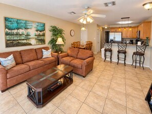 A living room with a sofa, coffee table, and a painting on the wall. There is a dining area with chairs and a table, and a kitchen with a refrigerator and cabinets.