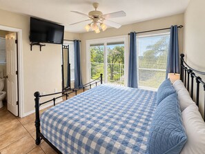 A bedroom with a bed, a television mounted on the wall, a ceiling fan, and a balcony with curtains.