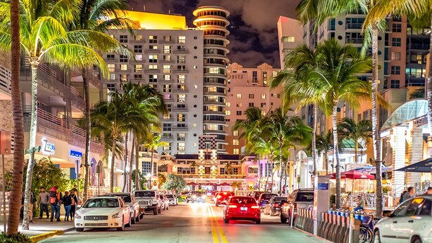 Una calle urbana nocturna con palmeras, edificios y coches aparcados.