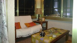 A living room with a sofa, a small table with a lamp, and a view of a cityscape through a window. - BALI-BENIDORM HOTEL FRONT APARTMENT (Finestrat)