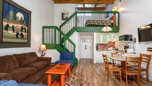 A cozy living room with a brown sofa, blue armchair, and a wooden coffee table. A green staircase leads to an upper level with a bed. The kitchen area is visible with a microwave, toaster, and a small dining table with chairs. - (A204) Loft Condominiums (Yosemite National Park)