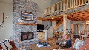 Ein gemütliches Wohnzimmer mit Kamin aus Stein, Holzmöbeln, einem Fernseher und einer Treppe zu einer oberen Ebene.