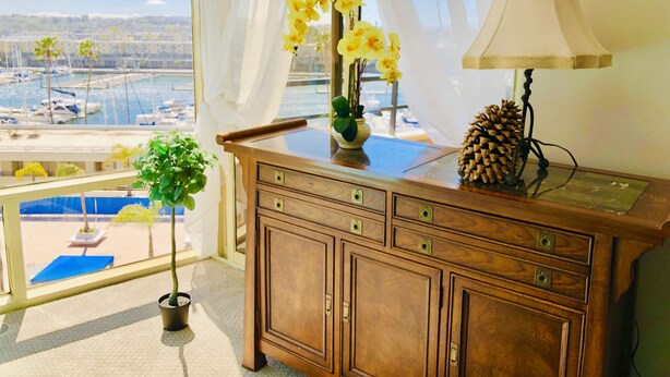 Ein Holztisch mit einer Vase voller gelber Blumen, einer Kiefernzapfe und einer Lampe. Im Hintergrund ist ein Blick auf einen Yachthafen und ein Gebäude zu sehen.