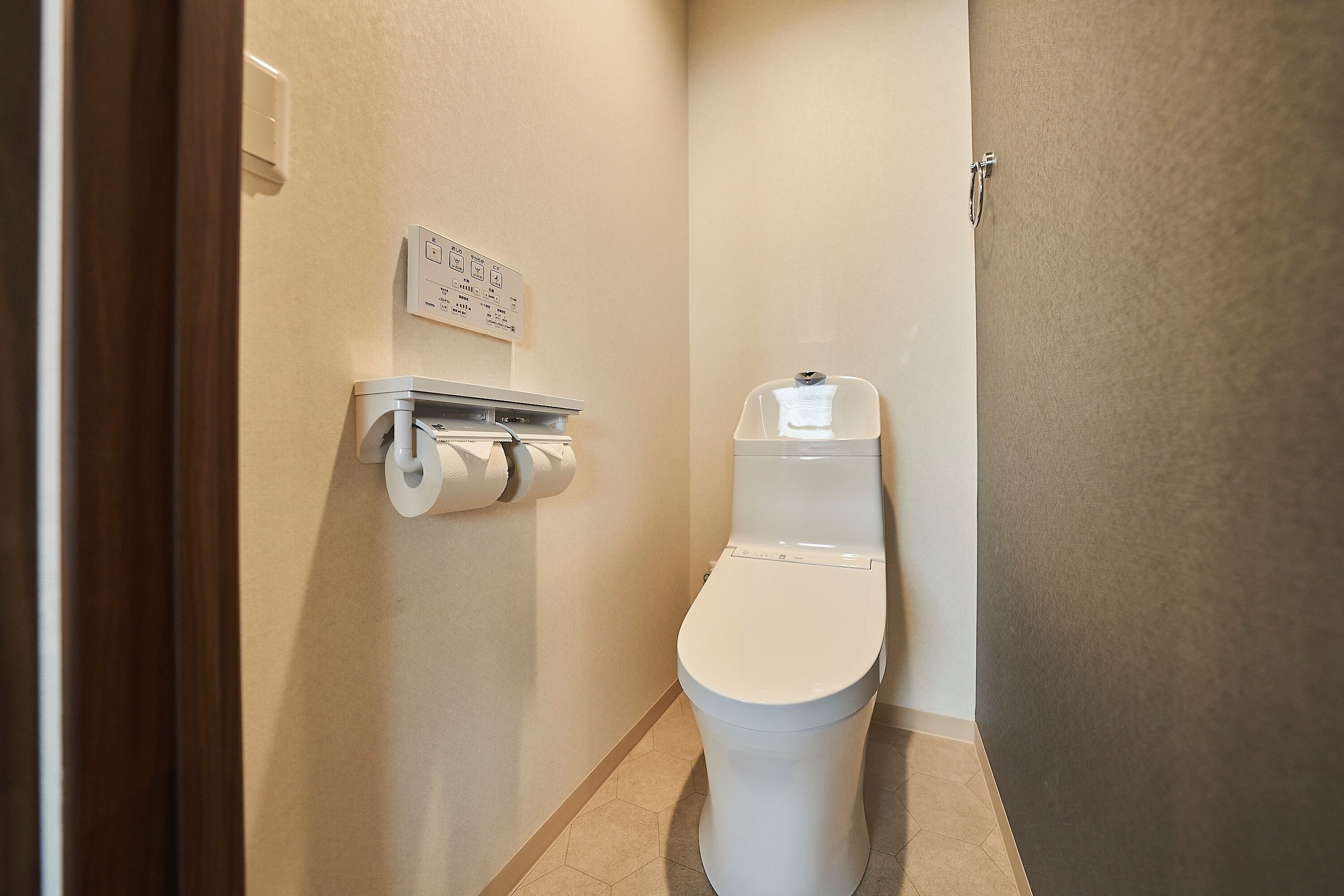 A small, clean bathroom with a toilet, toilet paper, and a wall-mounted flush control panel.