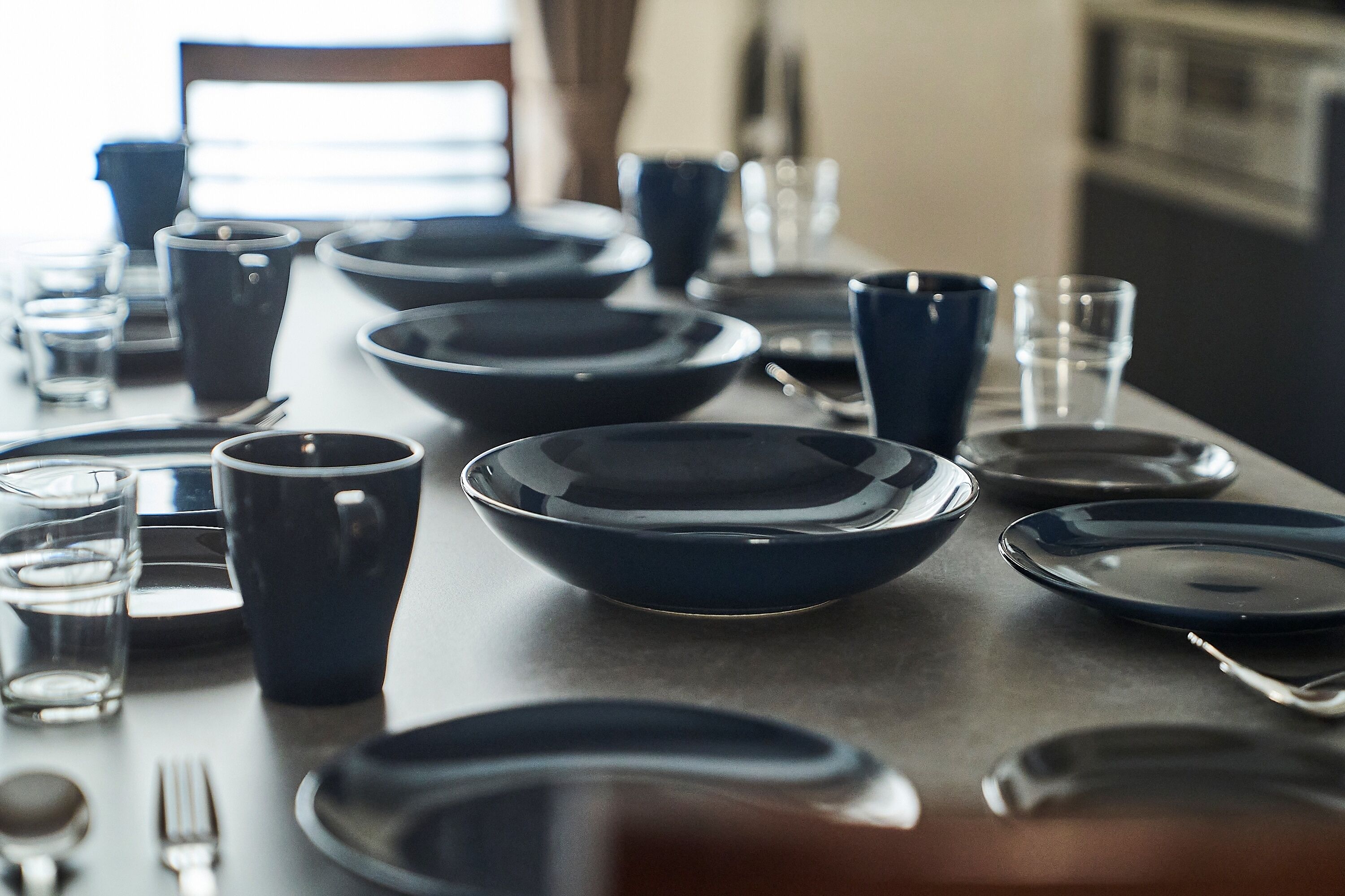 Une table à manger dressée avec de l’argenterie bleu foncé, comprenant des assiettes, des bols et des verres, le tout dans un style moderne et minimaliste.