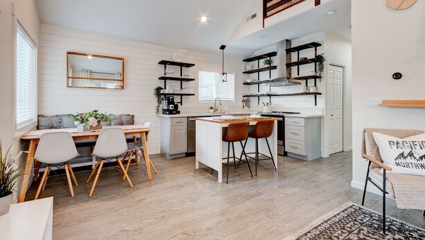 A modern kitchen with white cabinetry, wooden shelves, and a dining area with a bench and chairs.