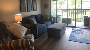 Uma sala de estar com um sofá azul escuro, uma poltrona listrada, um cobertor branco e fofo, e um tapete azul. Há três quadros na parede, um ventilador de teto e uma porta de vidro deslizante que dá para uma área externa com móveis de vime.