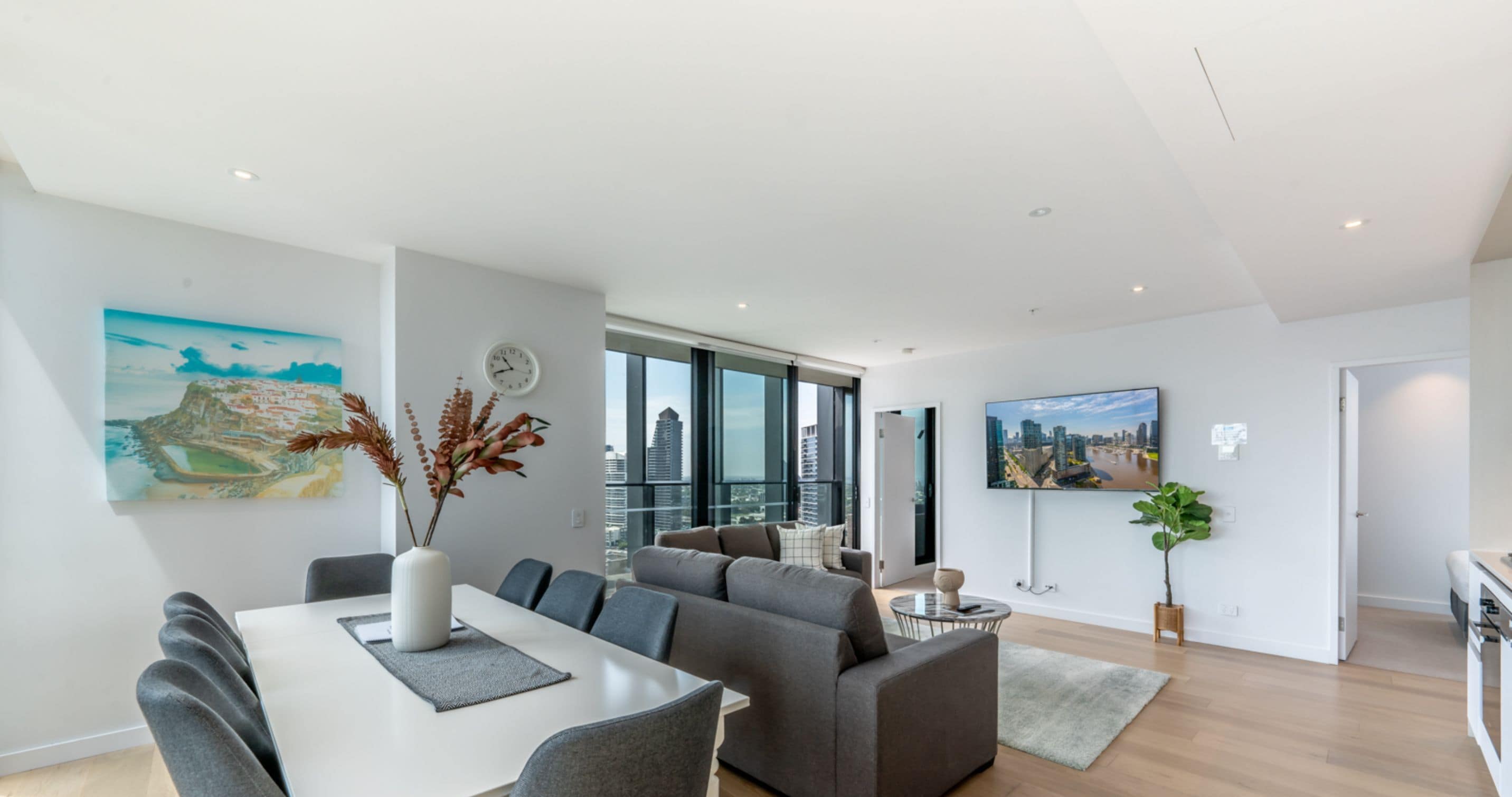 Ein modernes Wohnzimmer mit Essbereich, einem grauen Sofa, einem an der Wand montierten Fernseher und einem großen Fenster mit Blick auf die Stadt.
