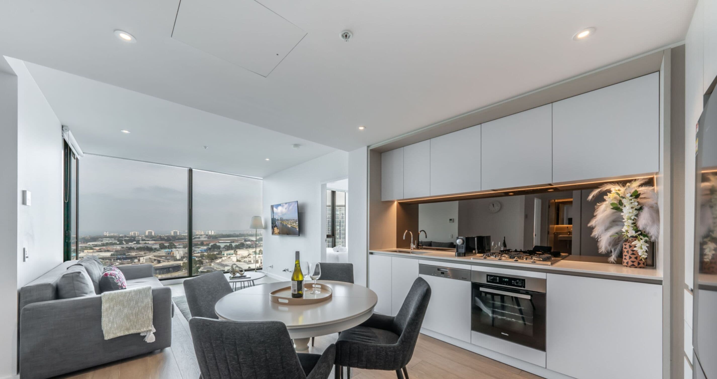 A modern apartment with a dining area, kitchen, and a large window offering a city view.