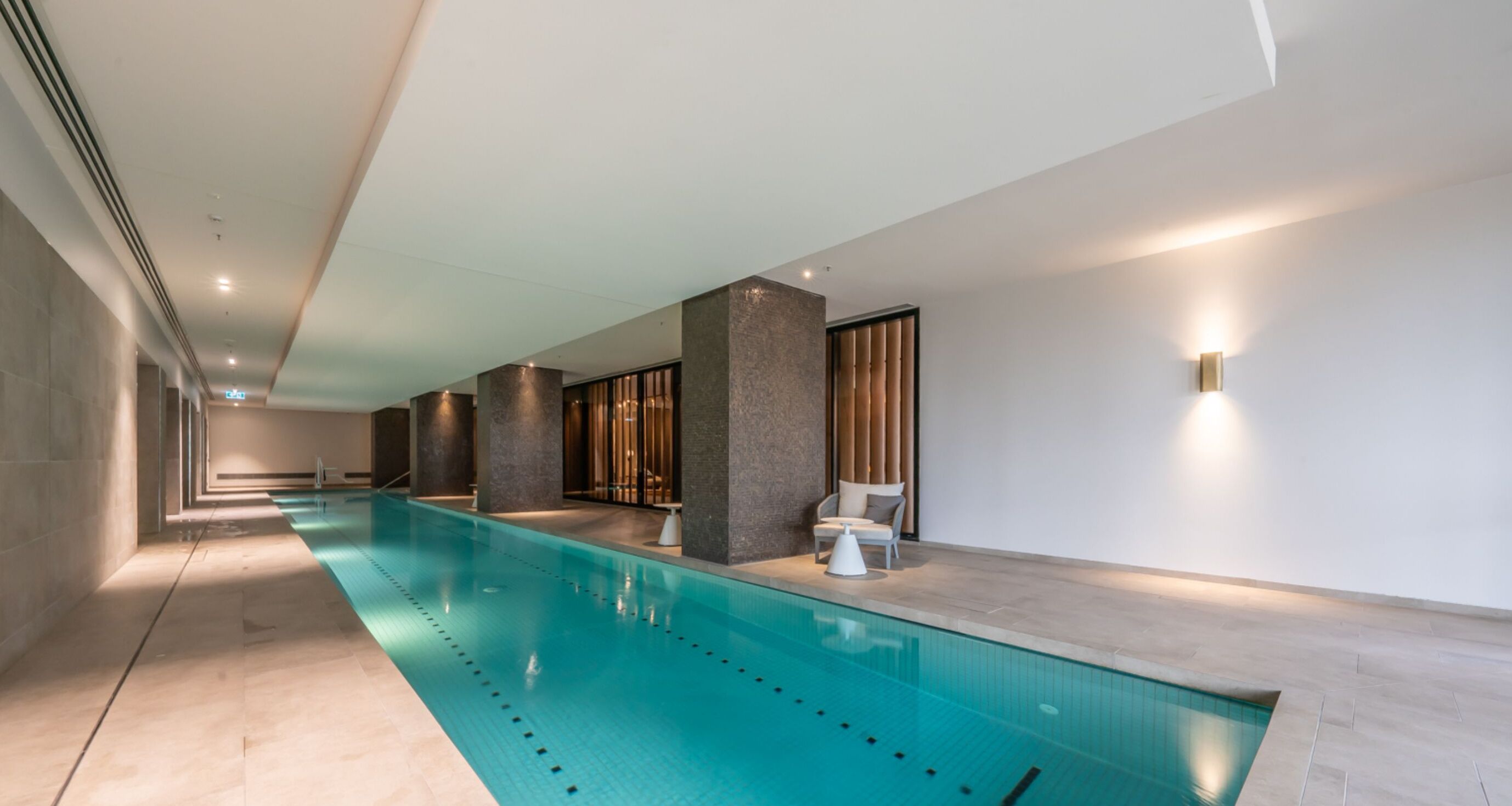 A modern indoor swimming pool with a long, narrow design, surrounded by a minimalist interior with neutral tones and a few seating areas.