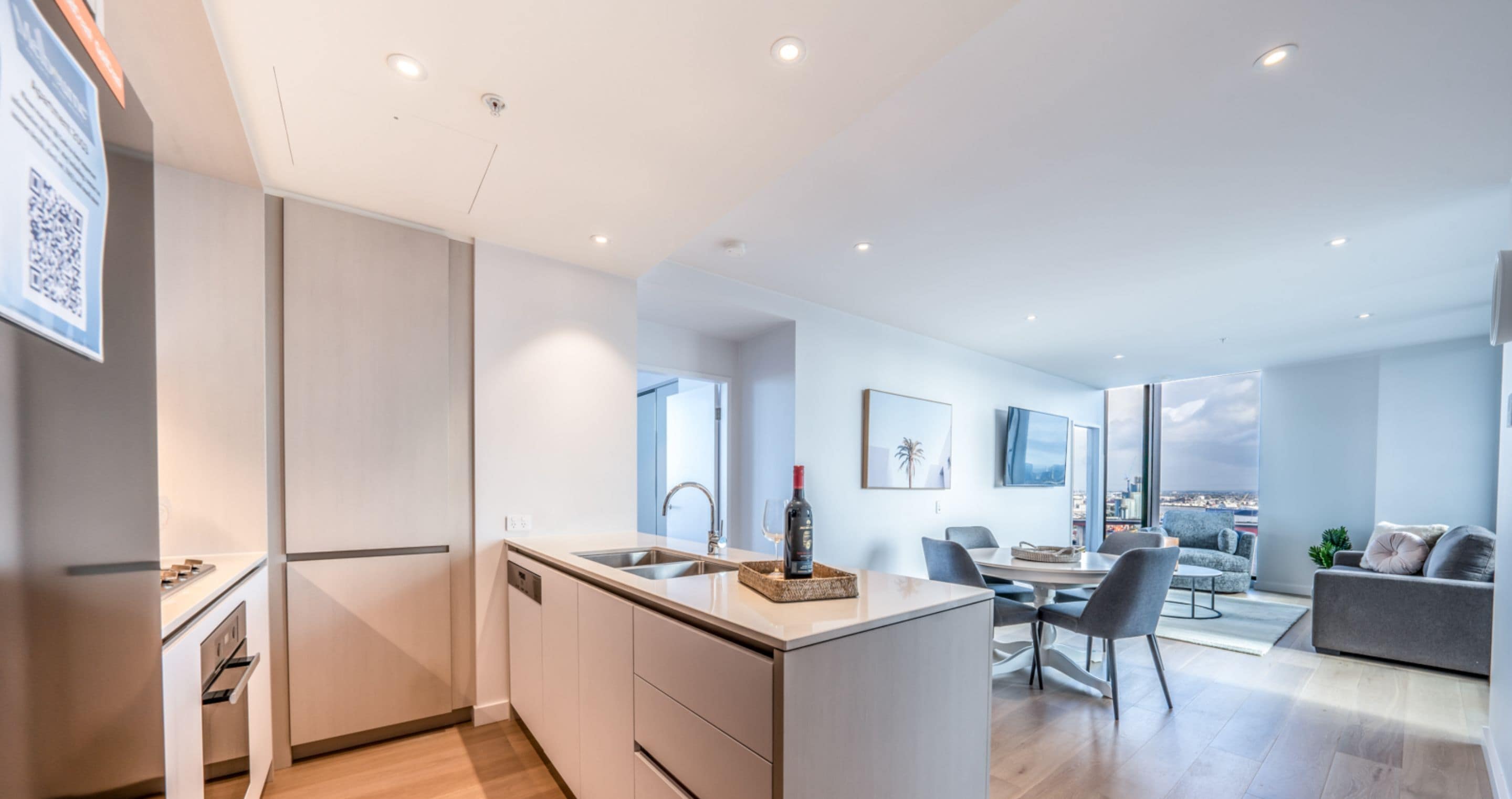 A modern kitchen with a central island, a dining area with a table and chairs, and a living space with a sofa and a city view.