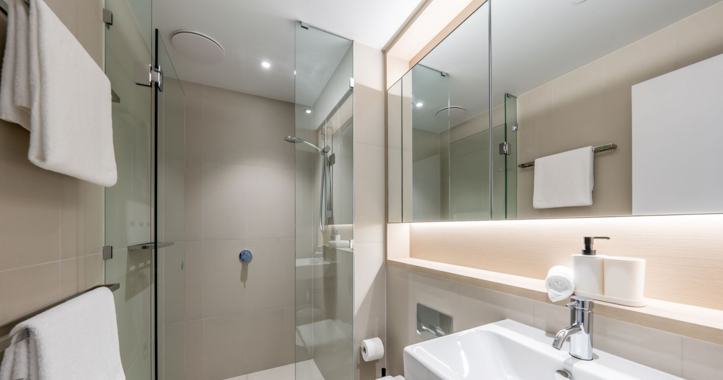 A modern bathroom with a glass-enclosed shower, a white sink with a soap dispenser, and a large mirror above the sink.