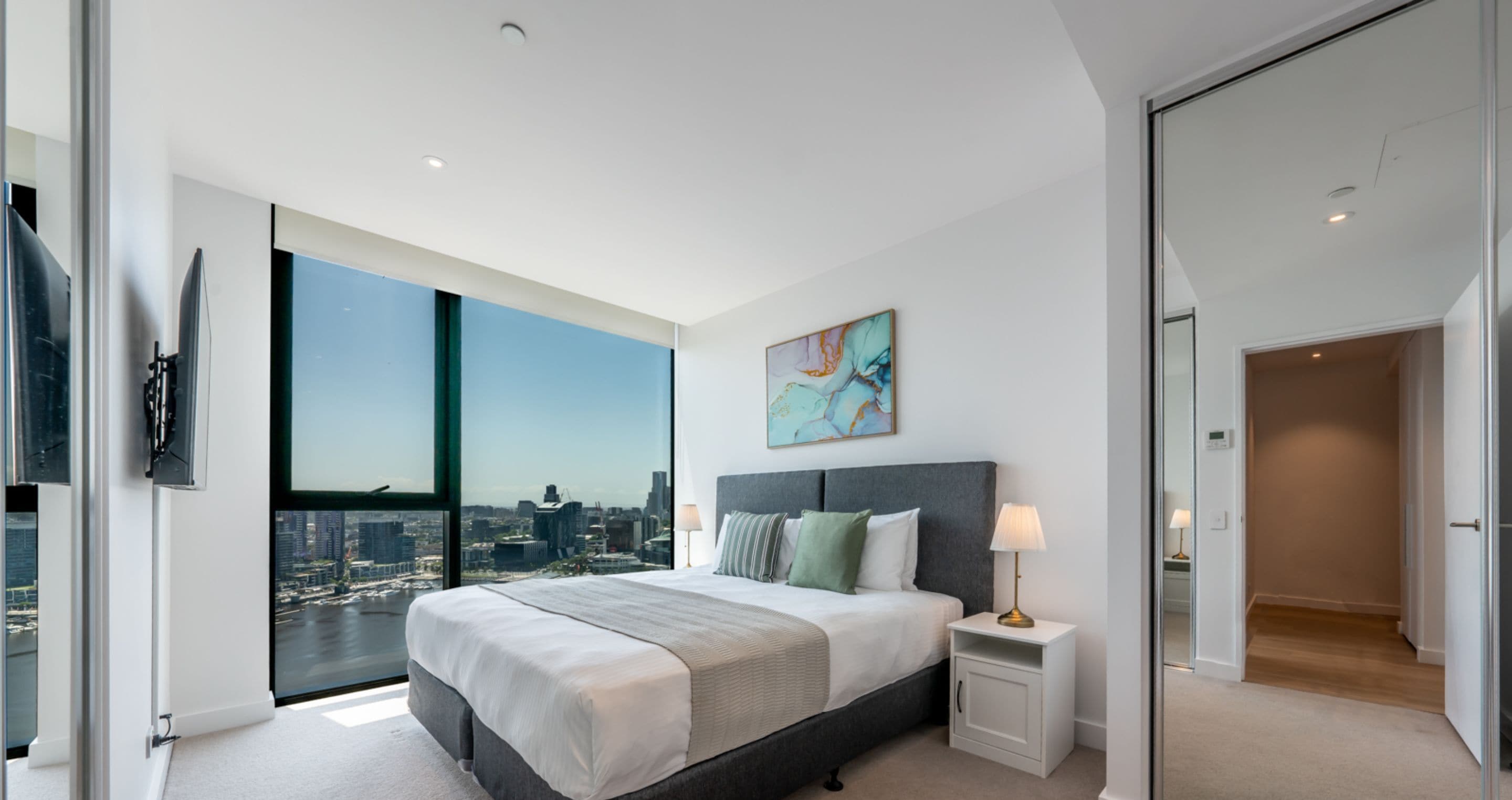 A modern bedroom with a large bed, a nightstand, a lamp, and a city view through floor-to-ceiling windows.
