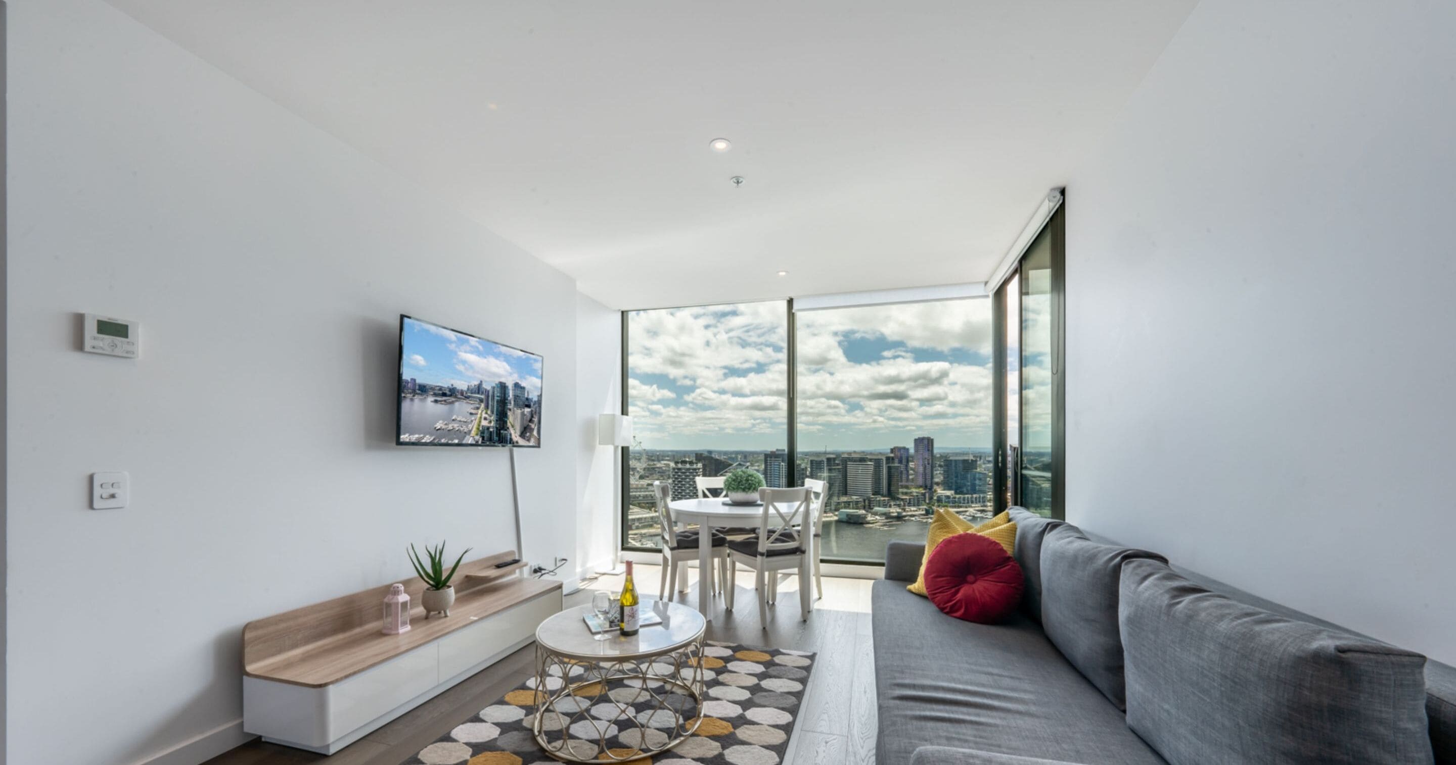 A modern living room with a large window, a flat-screen TV, a sofa, a coffee table, and a dining area with a view of the city.