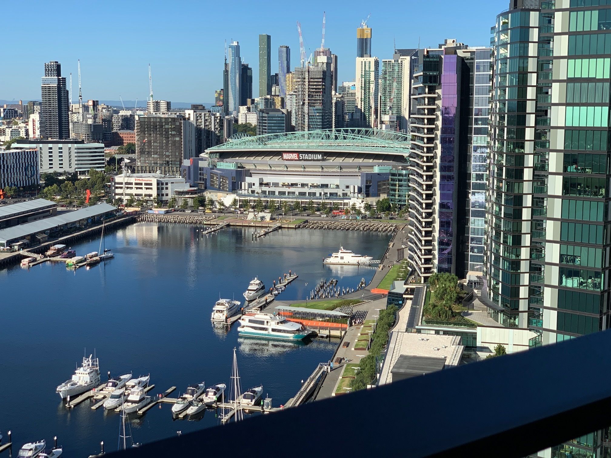 A marina with boats docked, a large stadium labeled "Marvel Stadium," and modern high-rise buildings in the background.