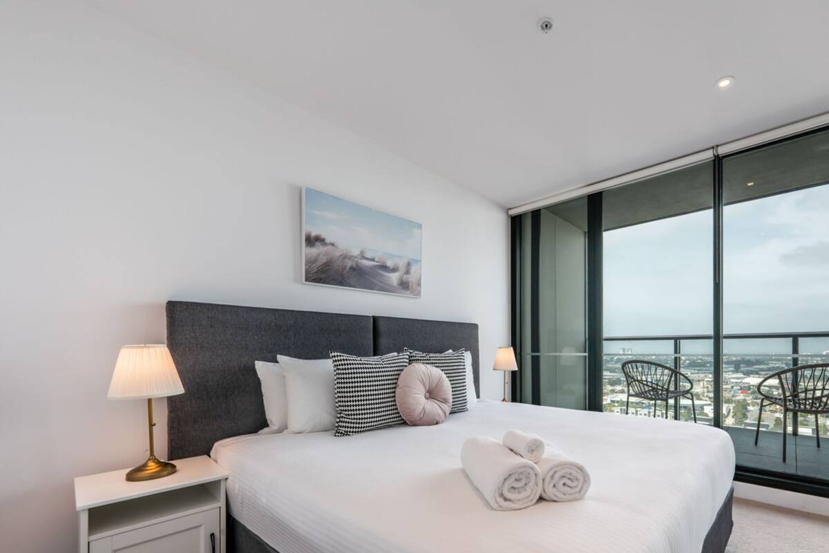 Een moderne hotelkamer met een groot bed, twee nachtkastjes, een stadszicht door de vloerhoogte ramen, en een stadslandschap schilderij boven het bed.