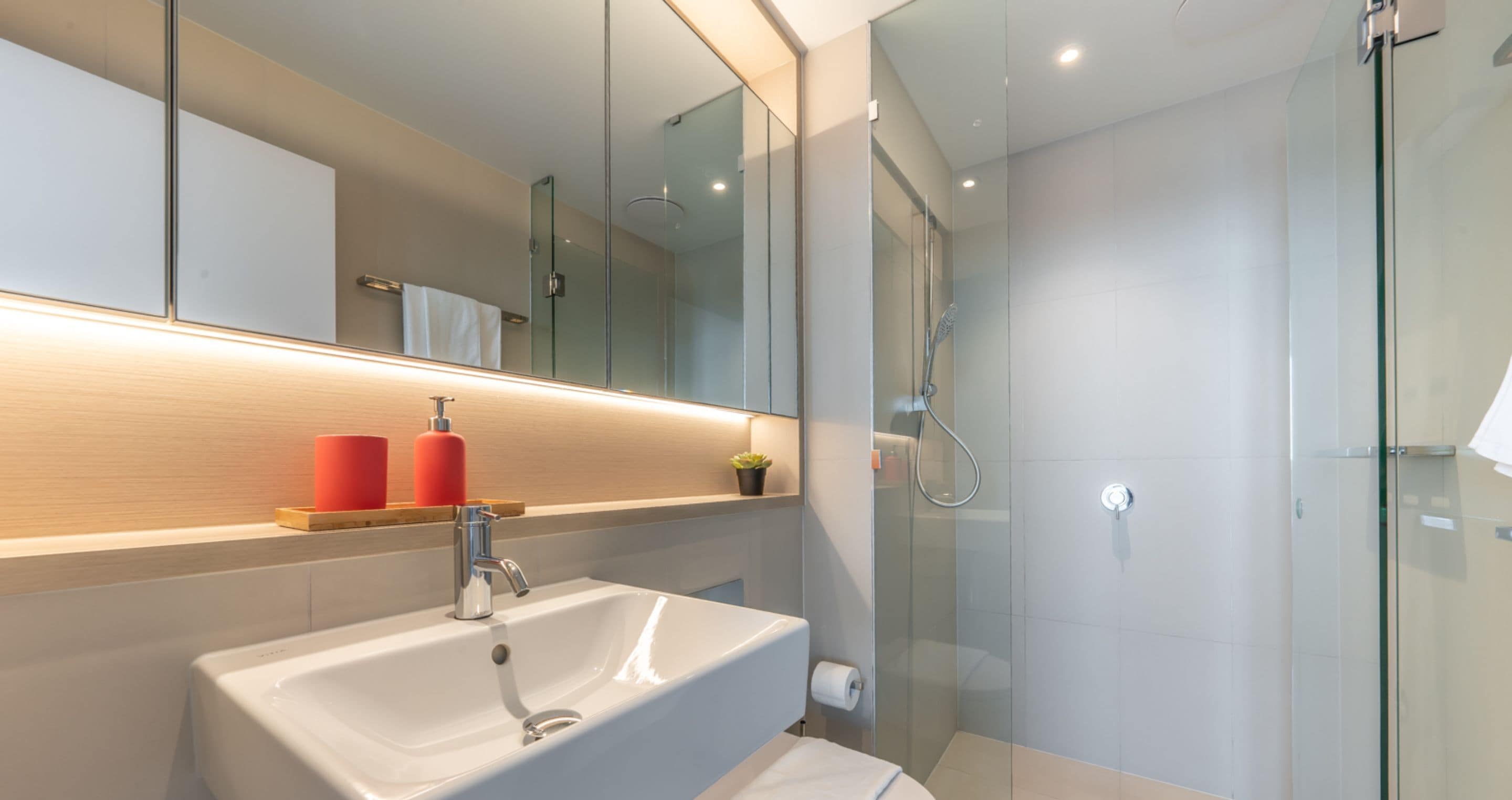 A modern bathroom with a glass shower enclosure, a white sink with a mirror above it, and a wooden shelf with two red bottles.