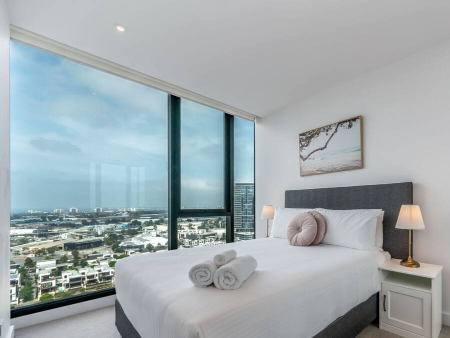 Una habitación de hotel moderna con un ventanal grande que ofrece vistas a la ciudad, una cama bien hecha con toallas, una mesita de noche con lámpara y una obra de arte enmarcada en la pared.