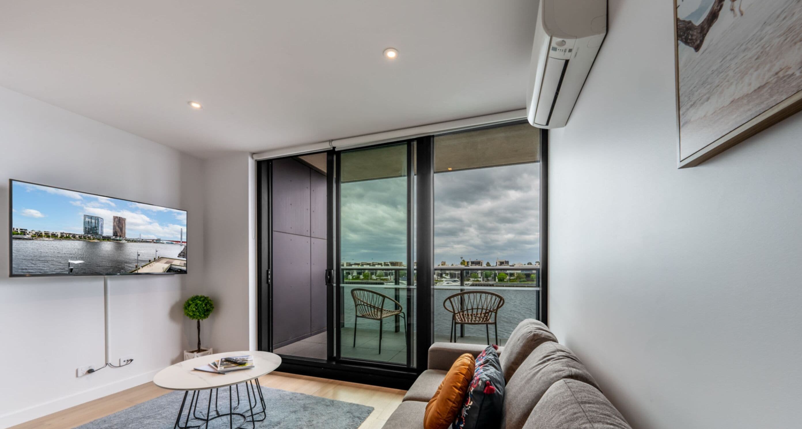 Uma sala de estar moderna com uma televisão grande, um sofá, uma mesa pequena e uma varanda com vista para a água e o horizonte da cidade.
