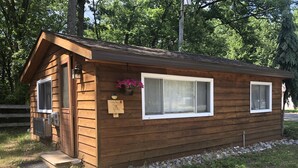 Eine kleine Holzhütte mit einer Tür und zwei Fenstern, umgeben von einem Kiesweg und viel Grün.
