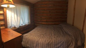 Ein Blockhaus-Zimmer mit einem Bett, einem Holznachttisch, einer Lampe und einem Fenster mit Vorhängen.