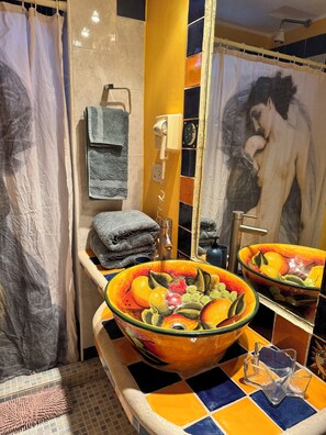 Une salle de bain avec un grand miroir, un lavabo orné d’un récipient aux couleurs vives, et un porte-serviettes.