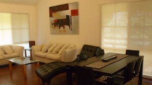 A living room with a sofa, a dining table with chairs, a painting on the wall, and a window with blinds. - Emada Venus Bay - a stone's throw from cafes and shops inVenus Bay (Venus Bay)