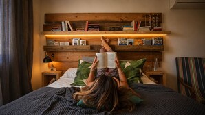Une personne allongée sur un lit, avec une bibliothèque au-dessus d’elle, est en train de lire un livre.