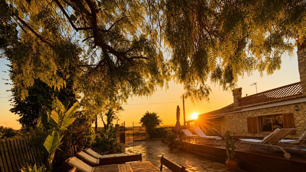 Une terrasse en bois avec des chaises longues, un bâtiment en pierre surmonté d’un toit en tuiles, et un coucher de soleil en arrière-plan.