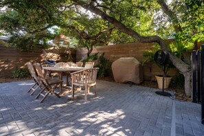 Une terrasse extérieure avec une table et des chaises en bois, un barbecue couvert et un ventilateur.