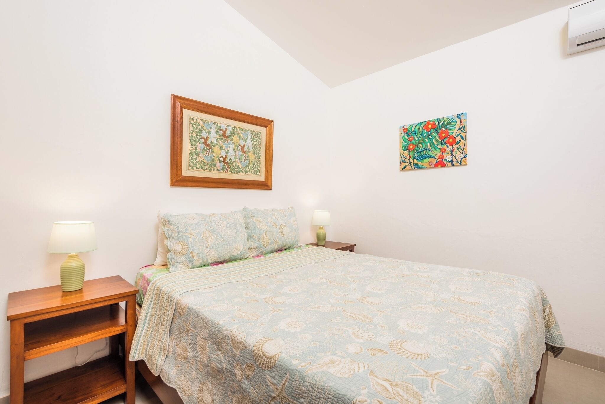 A bedroom with a bed, two bedside tables, a nightstand, a wall-mounted air conditioner, and two wall art pieces.