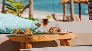 Una mesa de madera con un plato de comida, una bebida y un recipiente pequeño, sobre una playa con palmeras y una cabaña de paja al fondo.