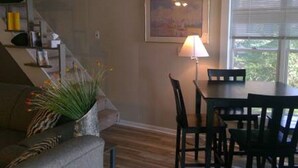 A living room with a sofa, a dining table with chairs, a lamp, a painting on the wall, and a staircase.