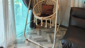 A room with a white swing chair, a brown cushion, a black sofa, and a view through a window with curtains. - Cozy house 1 BR, close to the beach (Tambon Choeng Thale)
