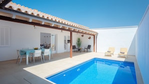 Et poolområde med en klar, blå pool, en overdækket terrasse med stole og et spisebord med stole.