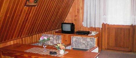A wooden interior with a dining table, chairs, a television, and a window with curtains.